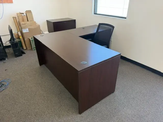 Contact Us L-shaped dark wood office desk with black mesh chair in a carpeted office room with boxes
