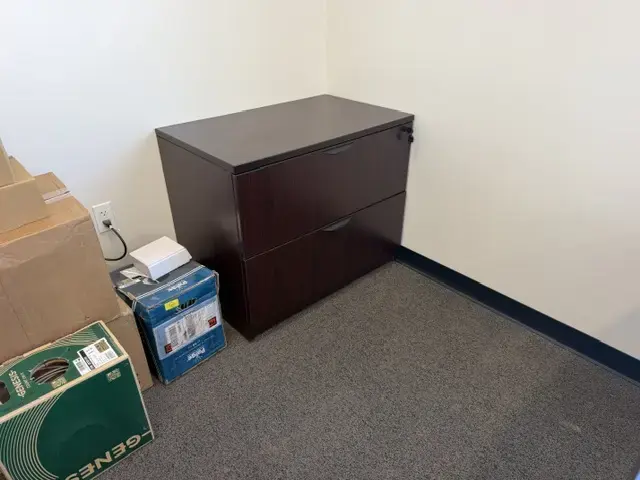 Contact Us Dark wood two-drawer lateral filing cabinet next to stacked cardboard boxes in an office corner