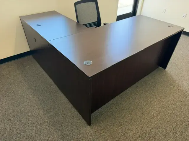 Contact Us Large L-shaped dark wood office desk with black mesh chair in a bright, carpeted office space