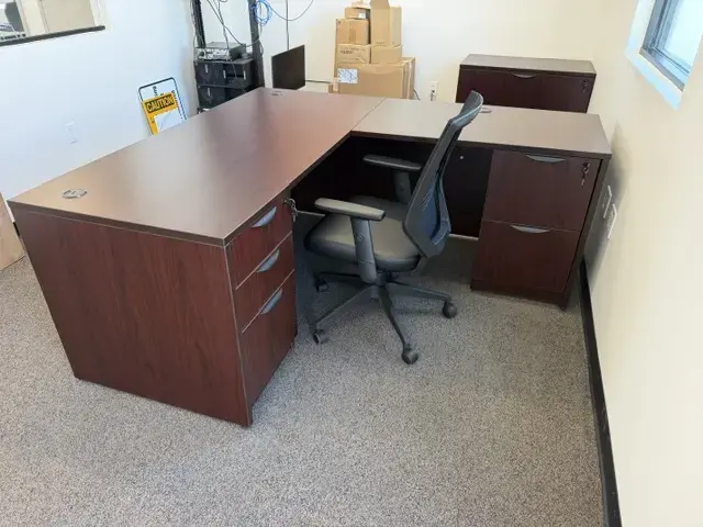 Contact Us Dark wood L-shaped office desk with three drawers and black mesh chair in a carpeted office with boxes