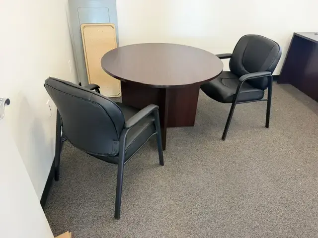 Contact Us Round dark wood meeting table with two black leather guest chairs on carpet in office corner