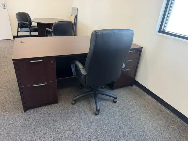 Contact Us Dark wood office desk with black leather chair and round meeting table with two chairs in carpeted office