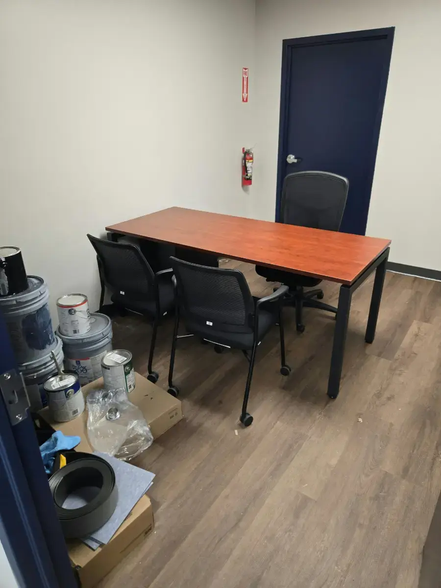 Let Us Help Build Your Next Workspace Small office with a wooden desk, three black chairs, and paint supplies stacked on the floor